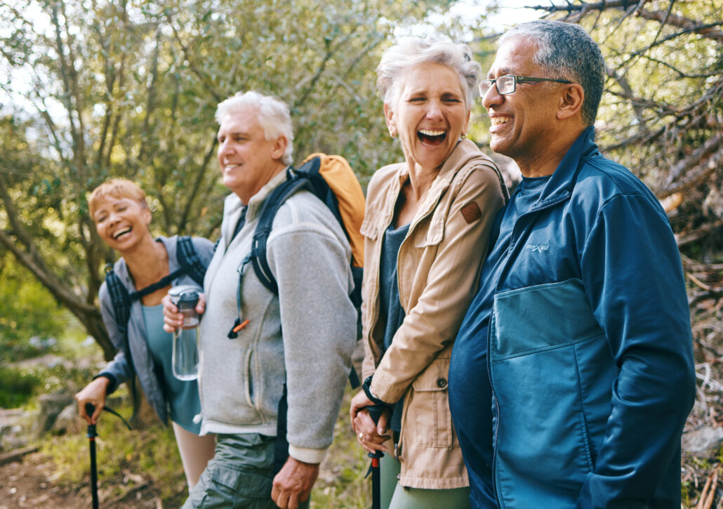 four older adults laugh together while hiking on a forest trail, enjoying an active and healthy lifestyle