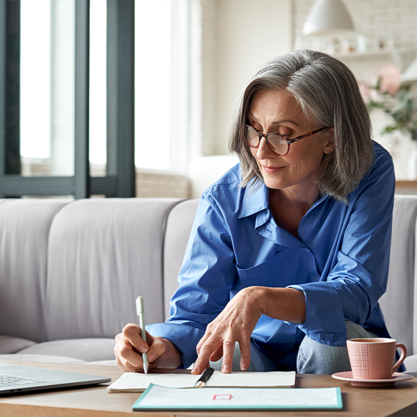mature woman with gray hair and glasses writes in a notebook while sitting on her couch with a cup of coffee nearby