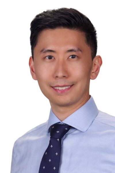 Professional headshot of male physician wearing a light blue shirt and navy patterned tie, smiling against a white background.