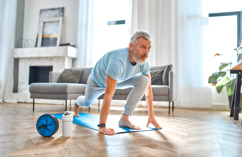 older man lunging on a yoga mat with exercise equipment beside him and a water bottle